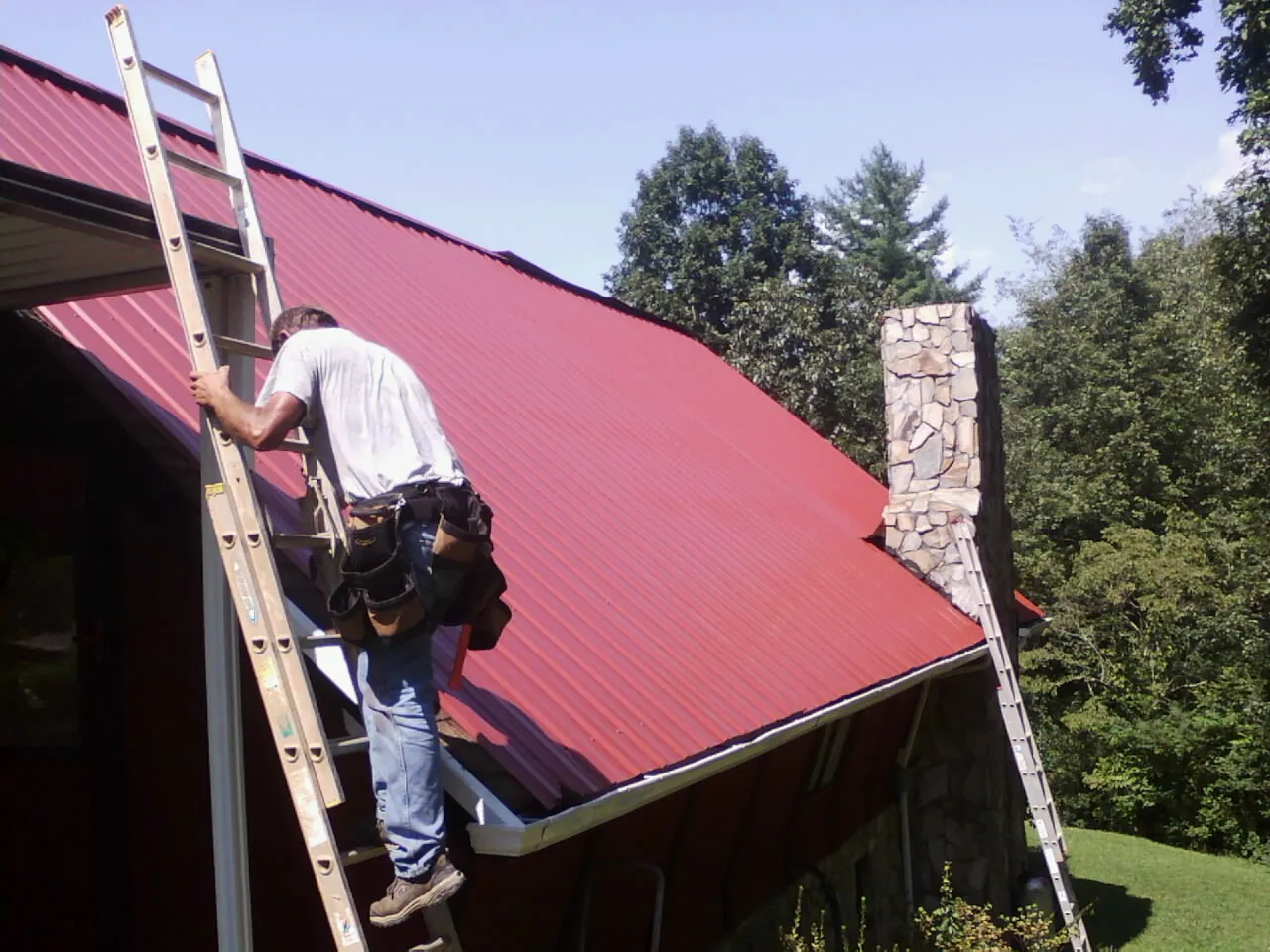 Roofer on a ladder beside a red metal roof and stone chimney — Soffit & Fascia Repair by Roofing Brothers in Moorpark